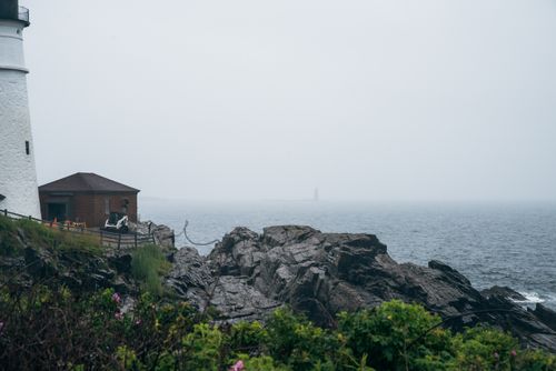 Free A misty seascape featuring a lighthouse, rocky shoreline, and distant fog-covered horizon. Stock Photo