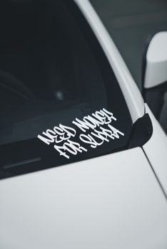 White car with humorous 'Need Money for Sushi' sticker on windshield, close-up view.