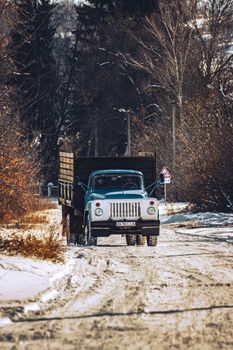 A vintage truck drives through a snowy, rustic road in winter, surrounded by bare trees.