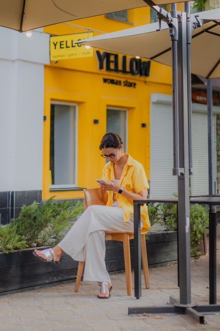 A woman wearing a yellow shirt sits outside a cafe, focusing on her smartphone.