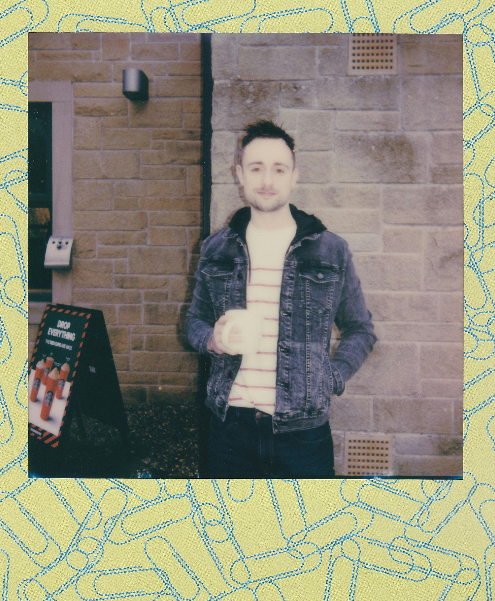 一名穿着牛仔夹克的男子靠在户外的砖墙上，拿着马克杯摆姿势