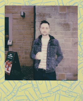 一名穿着牛仔夹克的男子靠在户外的砖墙上，拿着马克杯摆姿势