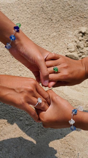 Three adults' hands adorned with colorful jewelry, signifying friendship and unity on a sandy beach.
