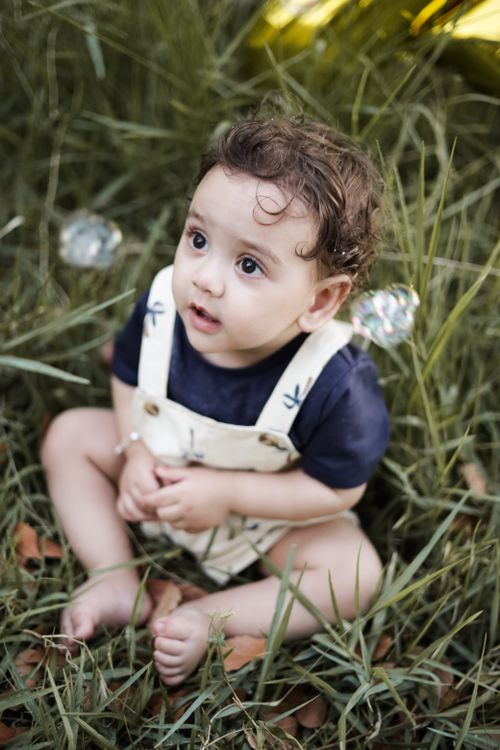 Free Cute baby in overalls sitting on grass, captivated by floating bubbles. Stock Photo