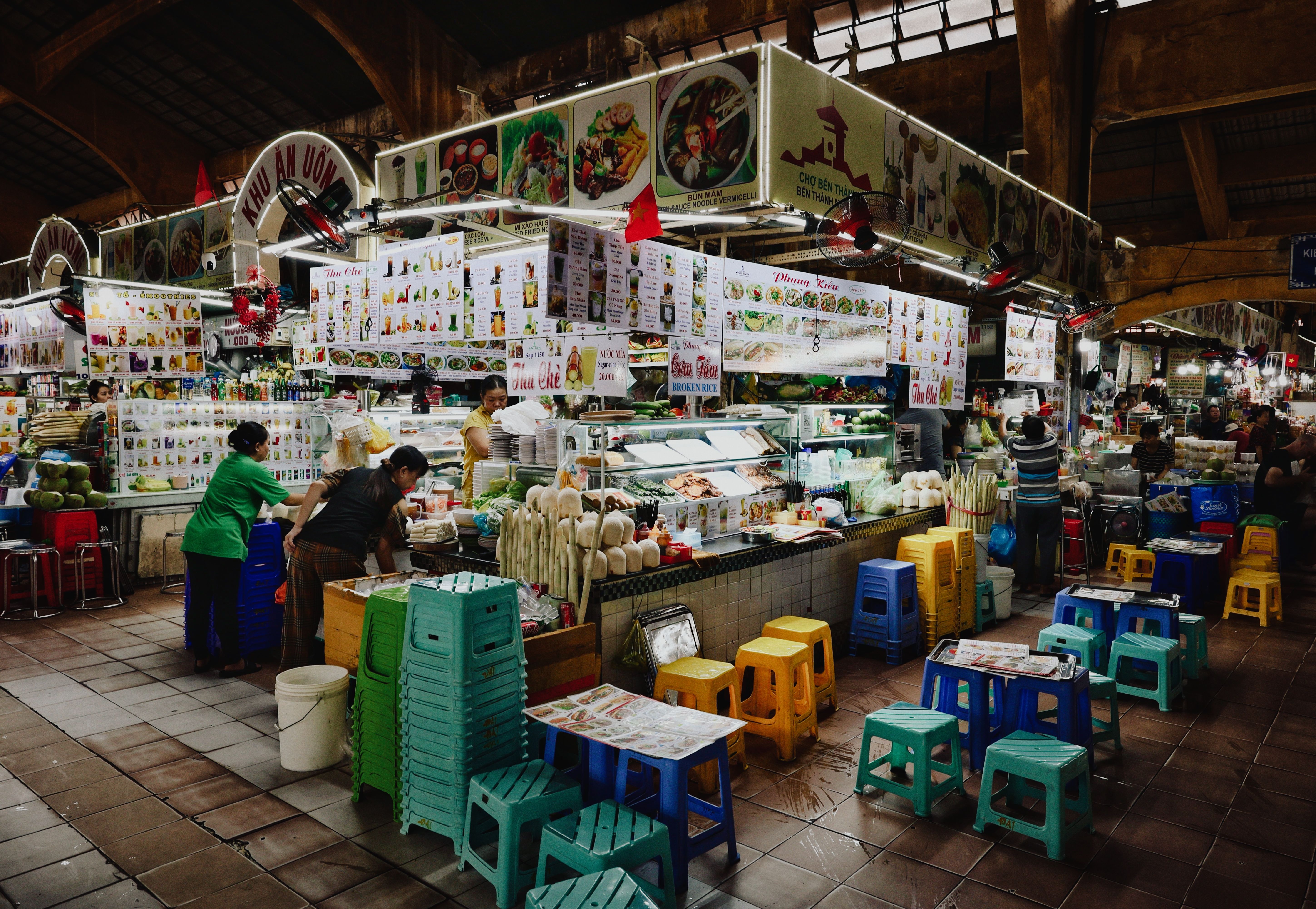 A vibrant food stall in a busy Vietnamese market with colorful seating and various food displays.