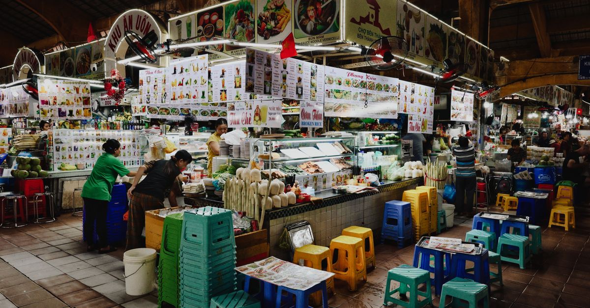 A vibrant food stall in a busy Vietnamese market with colorful seating and various food displays.