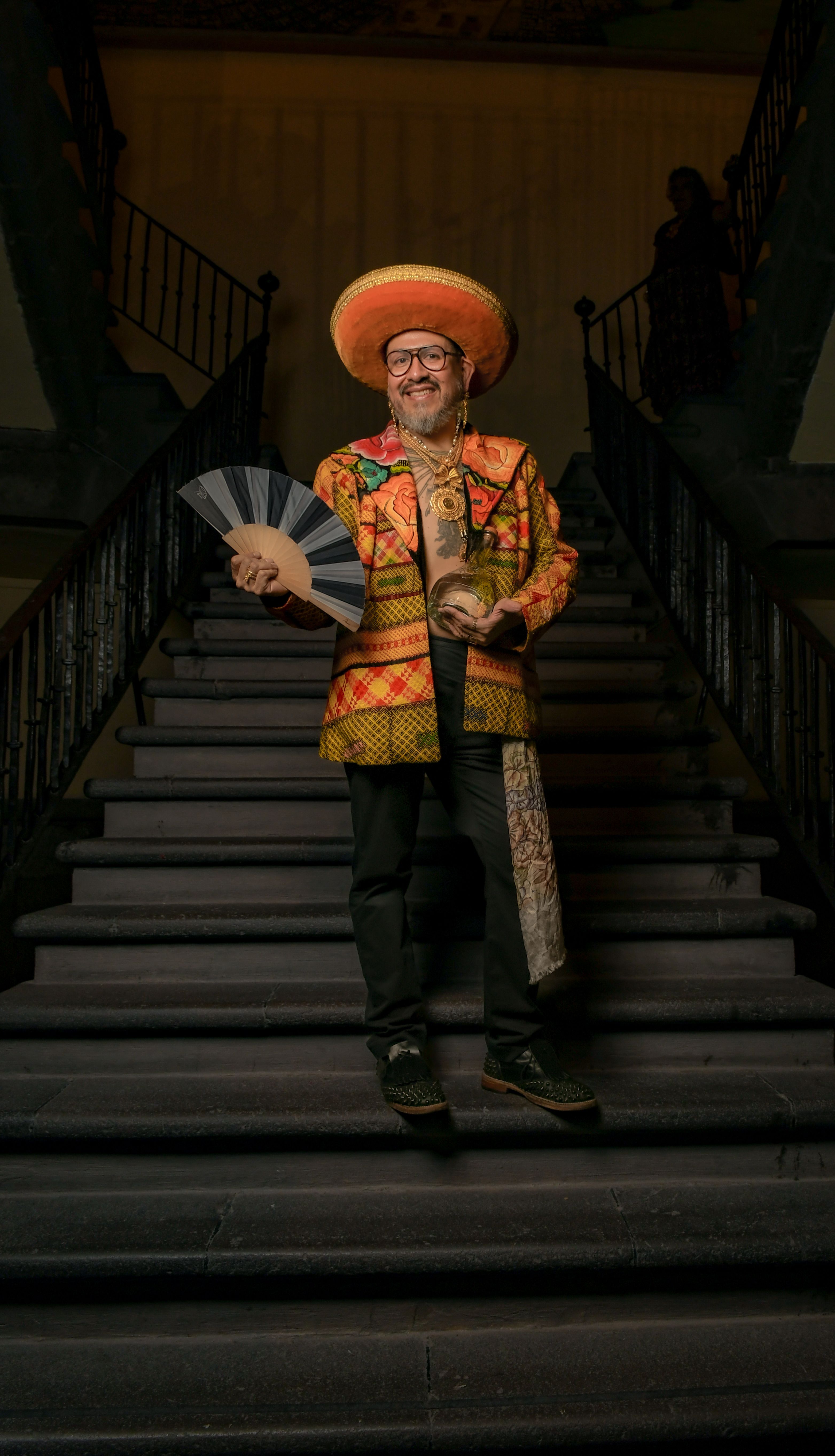 Smiling man in traditional attire holding fan on grand staircase indoors.