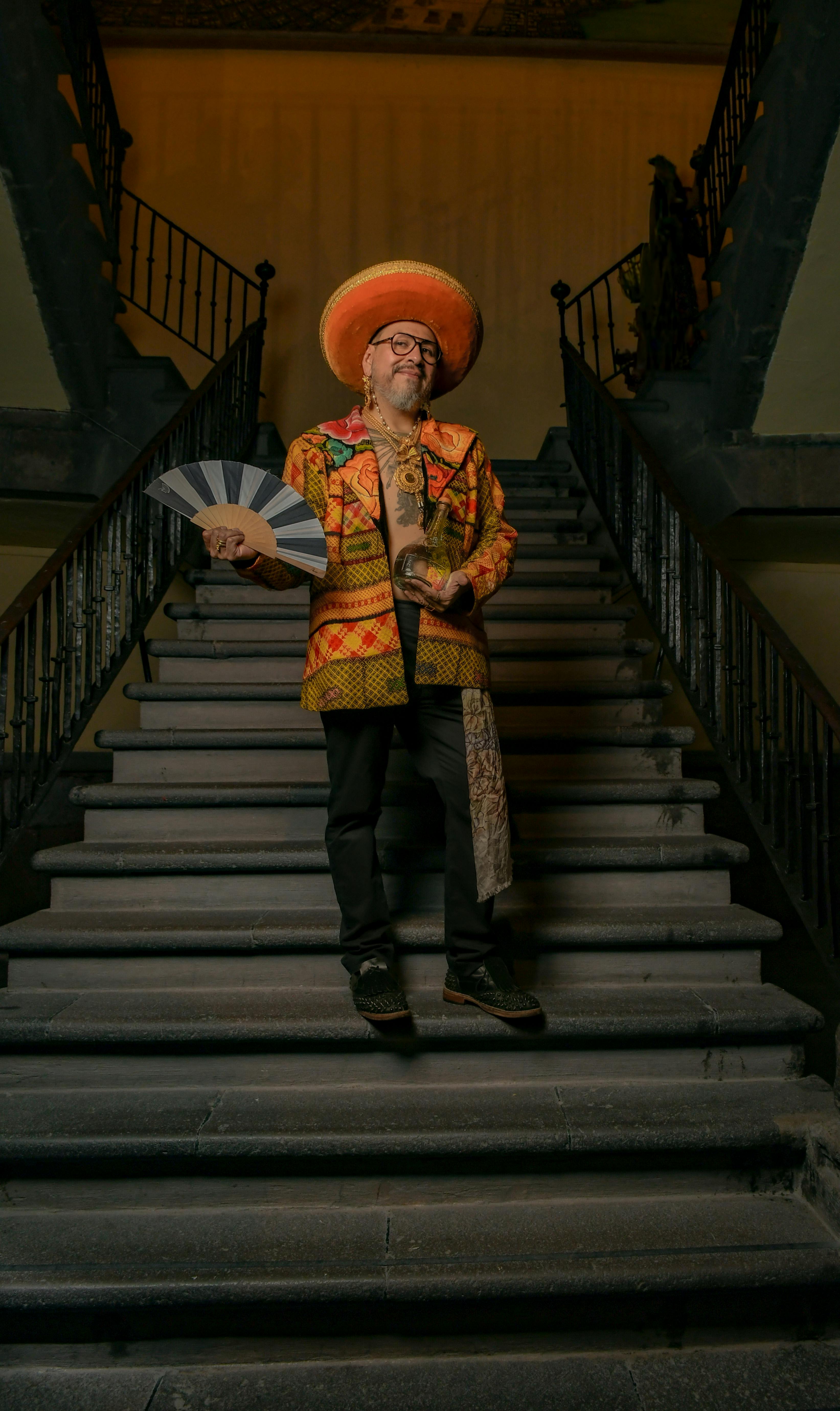 Man in vibrant outfit with fan and hat standing on a grand staircase indoors.