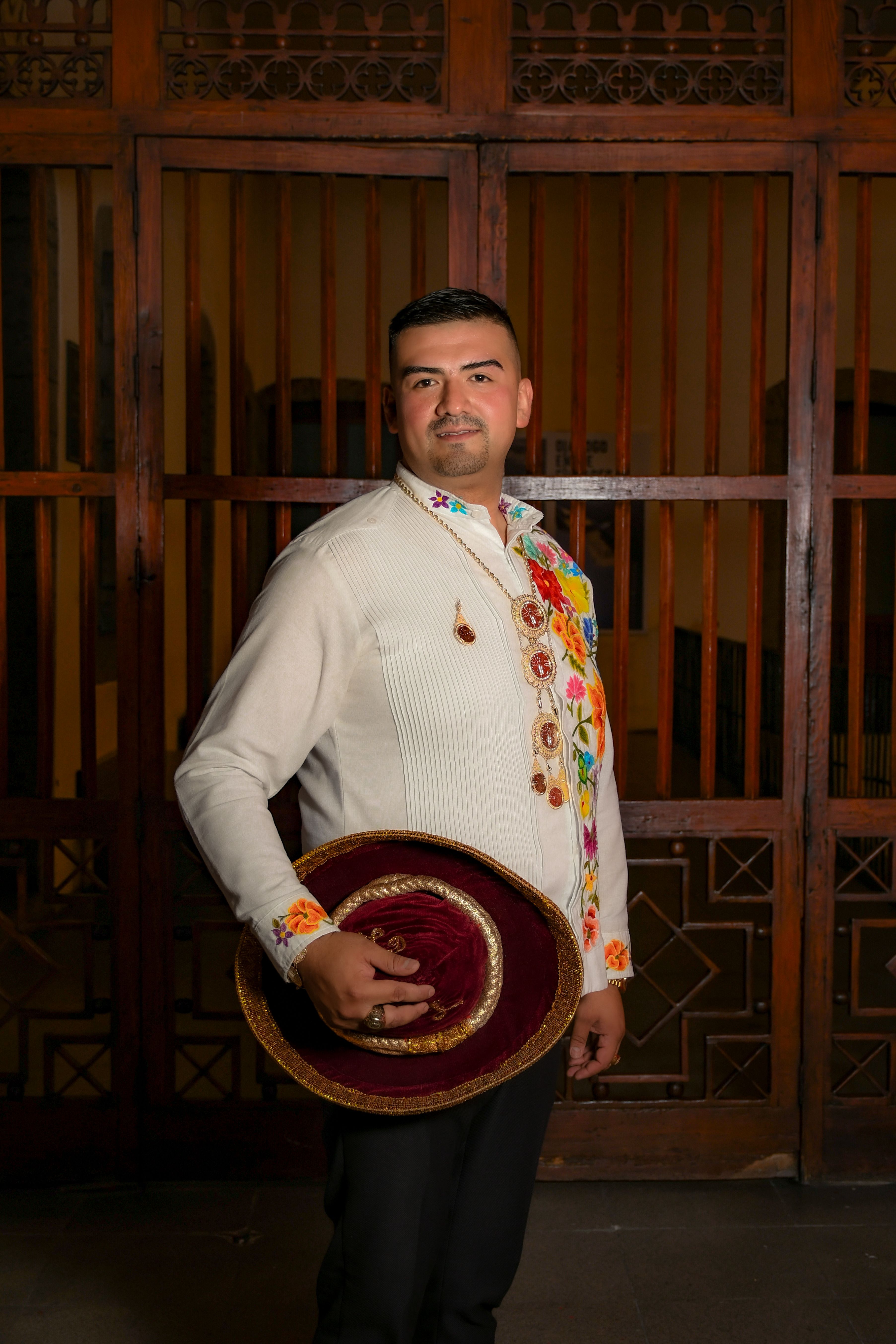 Man wearing traditional Mexican attire holding a decorative hat, posing indoors.