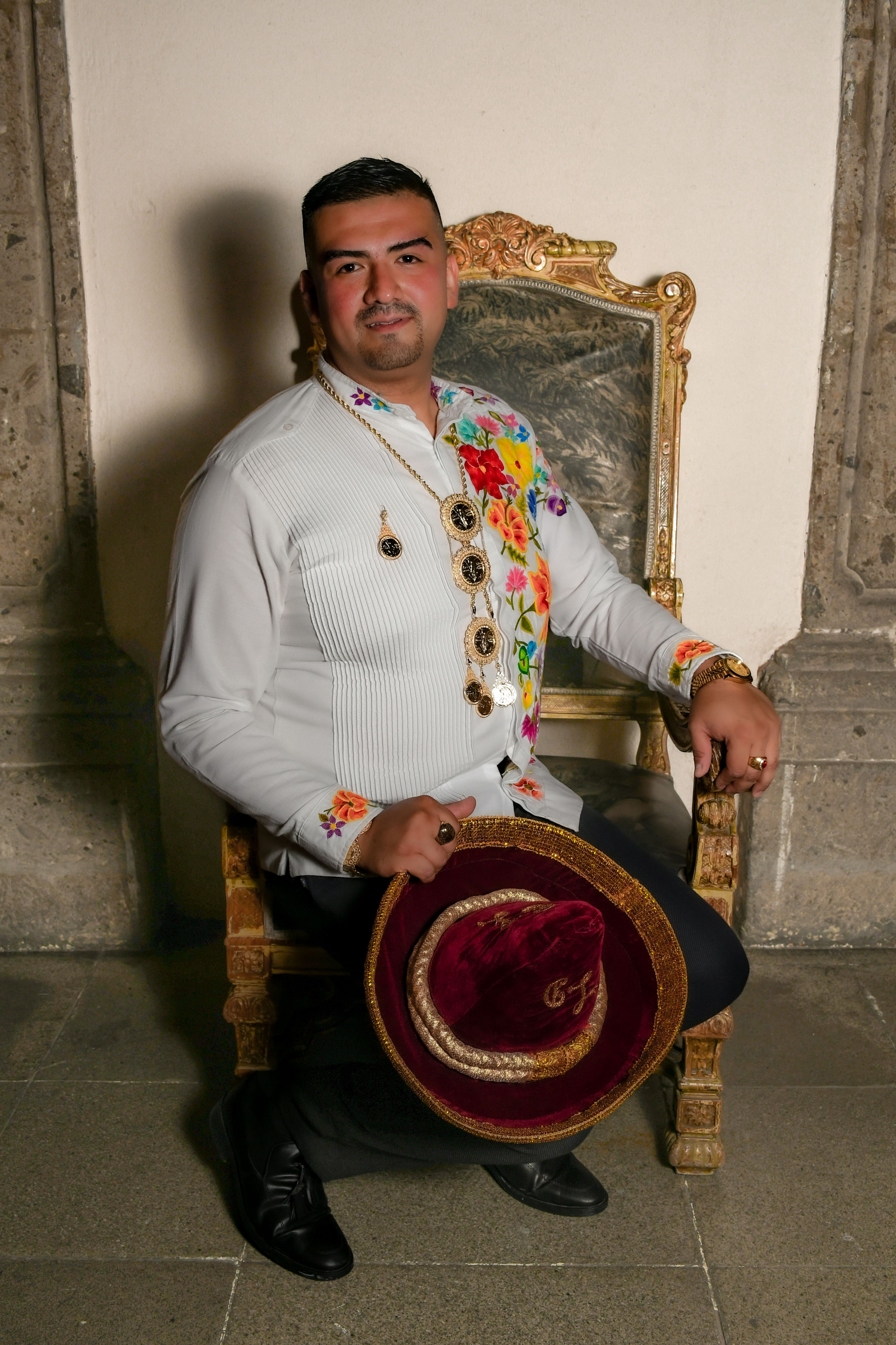 A man sits on an ornate chair wearing traditional embroidered clothing and holding a hat.
