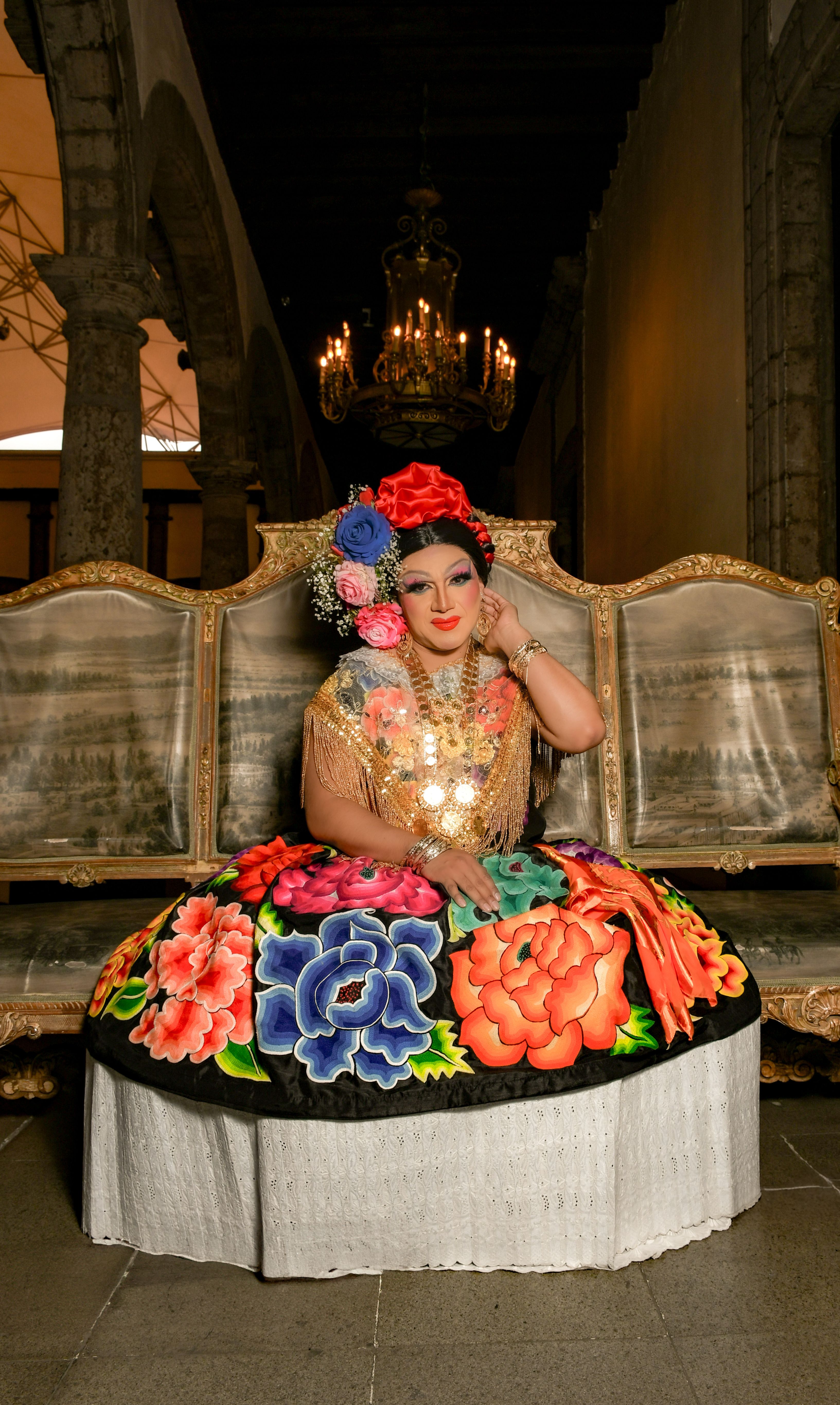 Person in vibrant cultural attire sitting on ornate bench indoors.