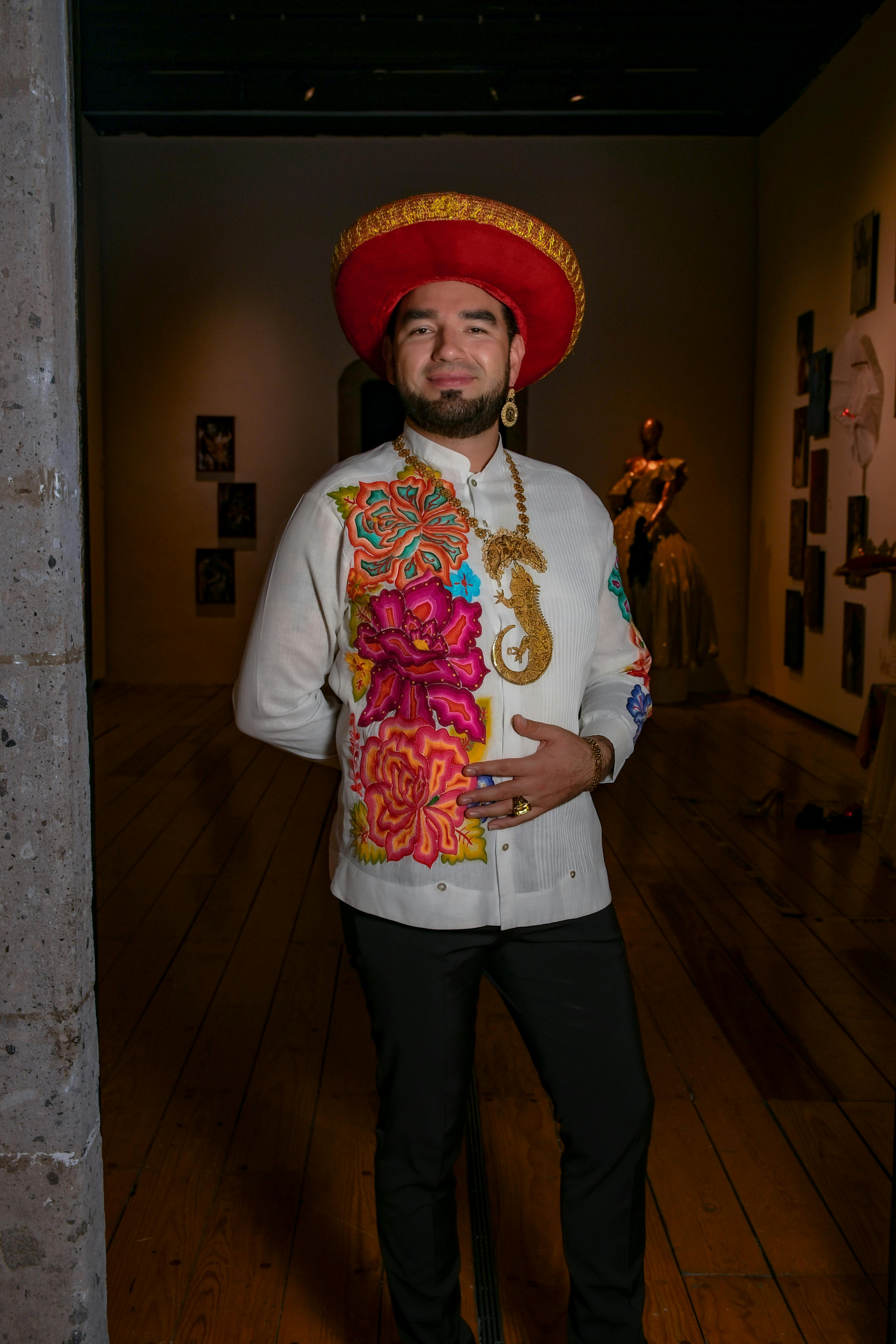 A man wearing a vibrant hat and shirt stands in a dimly lit art gallery.