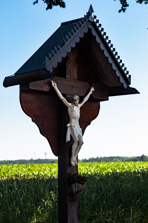 Free A rustic wayside cross with a depiction of Christ in a vibrant, lush outdoor setting. Stock Photo