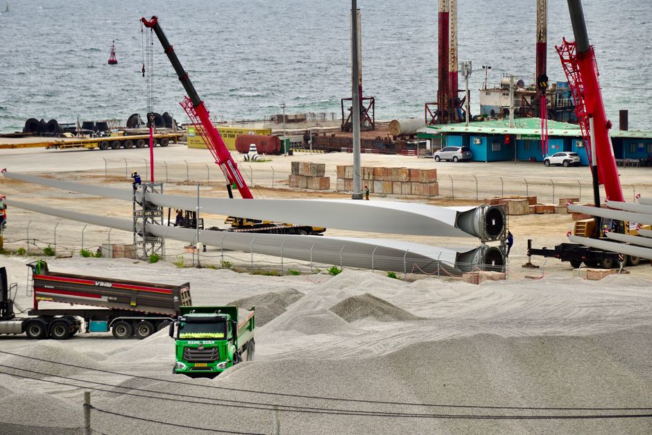 Construction site featuring wind turbine blades, cranes, and trucks by the seaside, showcasing industrial activity.