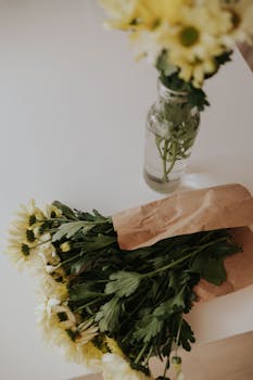 A bouquet of fresh daisies wrapped in brown paper next to a vase on a white table.