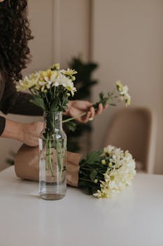 A person arranges a bouquet of white and yellow daisies in a glass vase indoors.