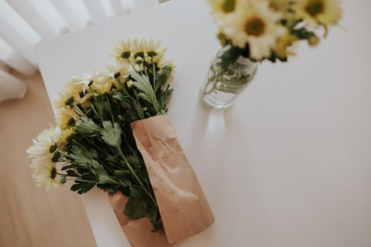 Flat lay of yellow chrysanthemums in paper wrap and vase on a clean white table.