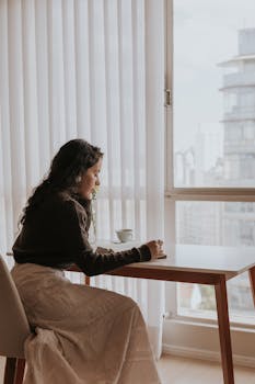 A woman sits by a window with a city view, writing in a notebook, creating a serene atmosphere.