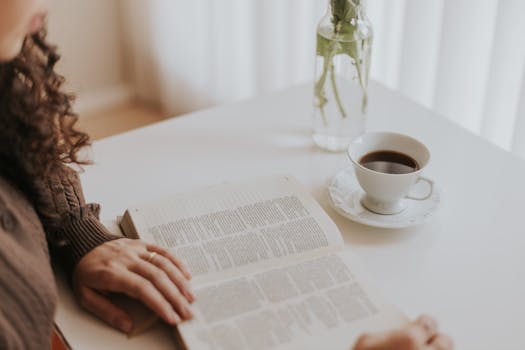 Woman reading a book at a white table with coffee and a vase, creating a cozy moment.