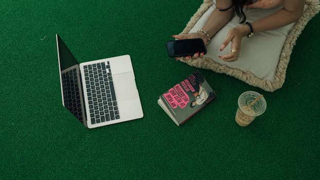 Woman relaxing with laptop, book, and iced coffee on green grass.