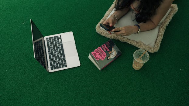 Woman using a smartphone by a laptop, book, and iced coffee on green turf.