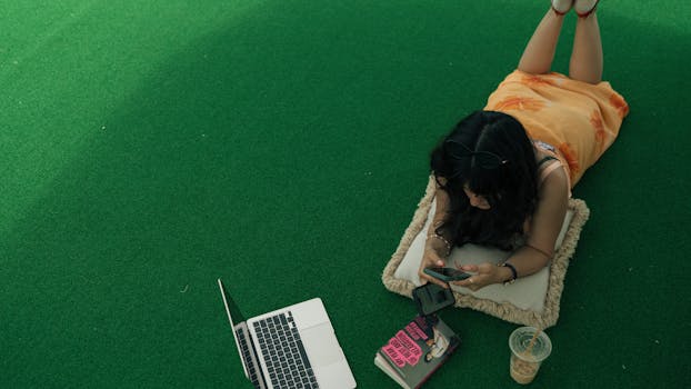 Woman lying on a cushion outdoors using phone, with laptop and book nearby.
