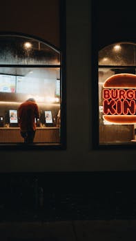 Moody nighttime view of a Burger King with glowing neon signs and a person at the counter.