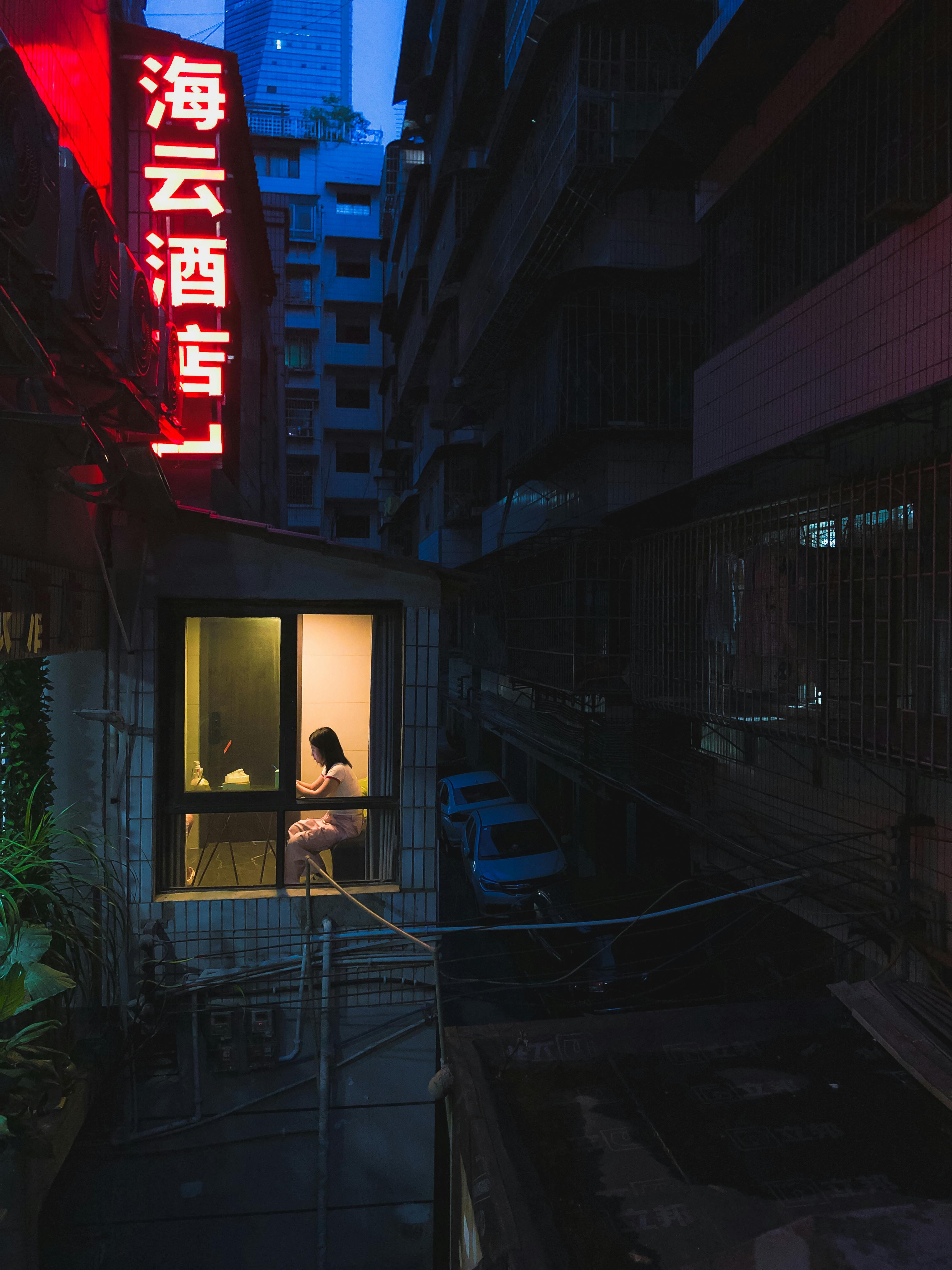 A moody urban evening with a neon sign illuminating a lone figure in a window.