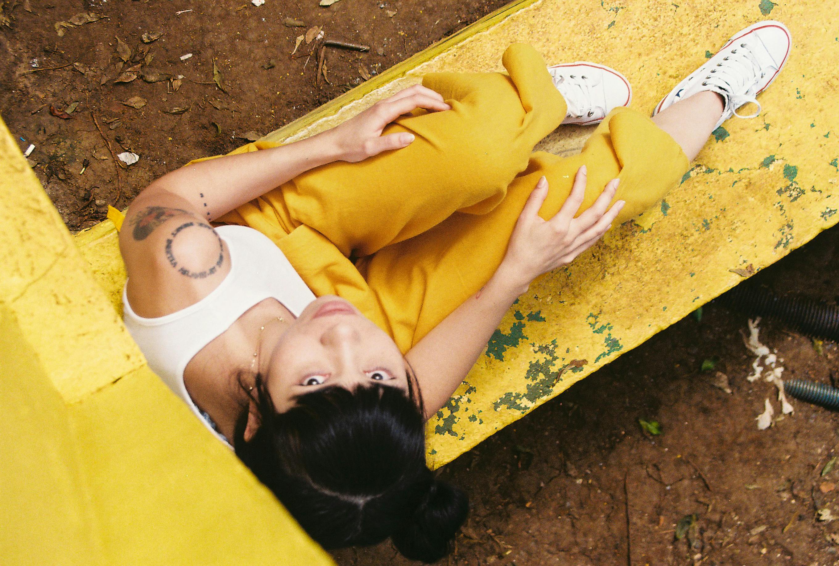 Young woman in yellow pants relaxing on a bench outdoors, shot from above, showcasing a casual and vibrant style.