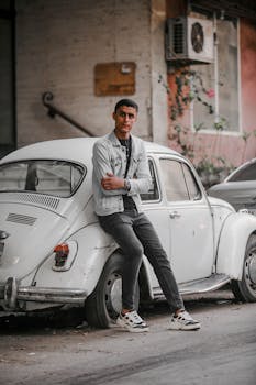 一个穿着休闲装的年轻人靠在一辆停在户外的白色经典大众甲壳虫上
