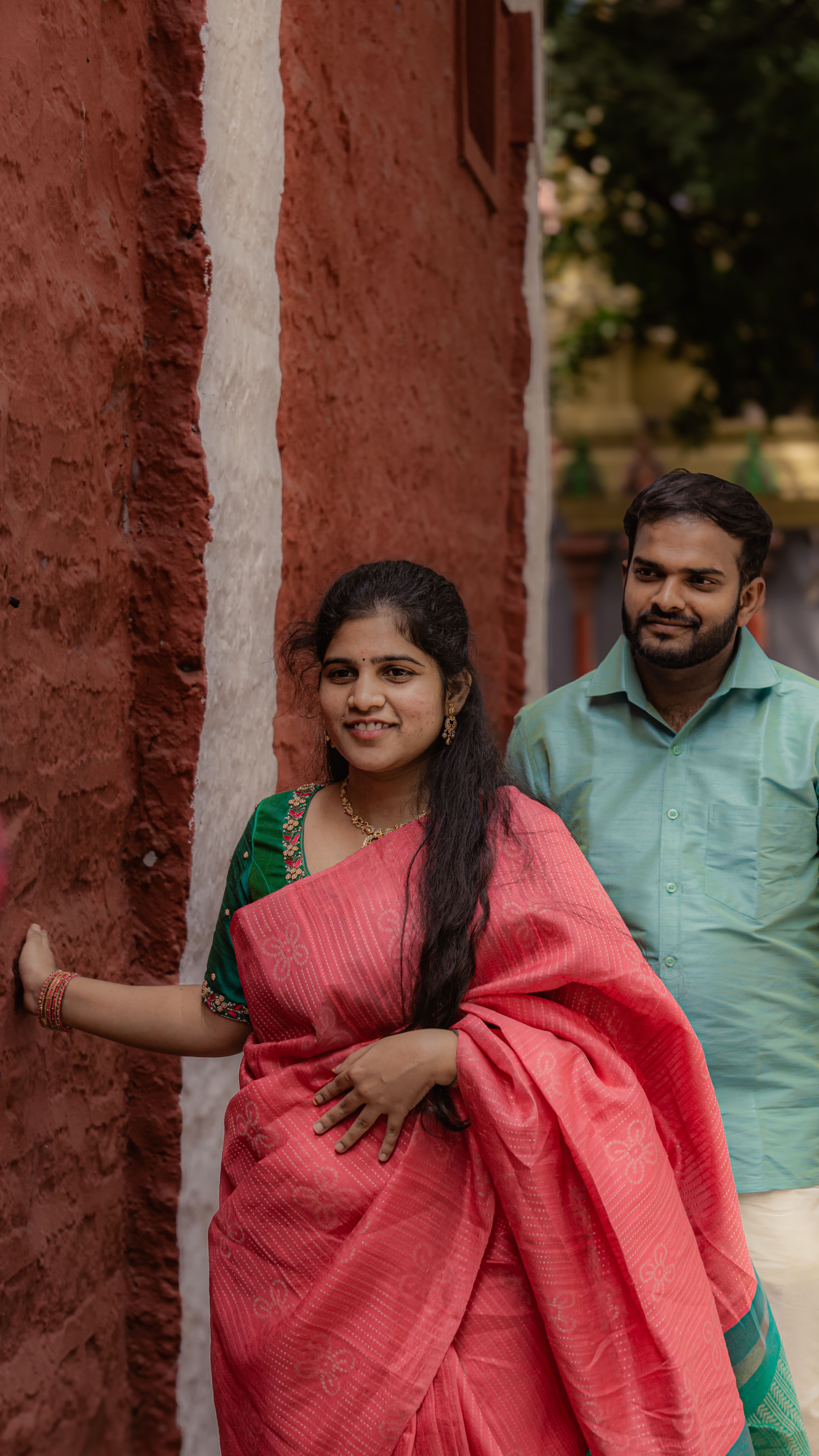 Happy couple in traditional attire walking along a rustic red wall outdoors.