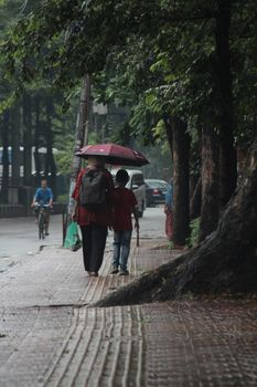 A mother and child walk under a red umbrella on a wet city sidewalk, surrounded by greenery.