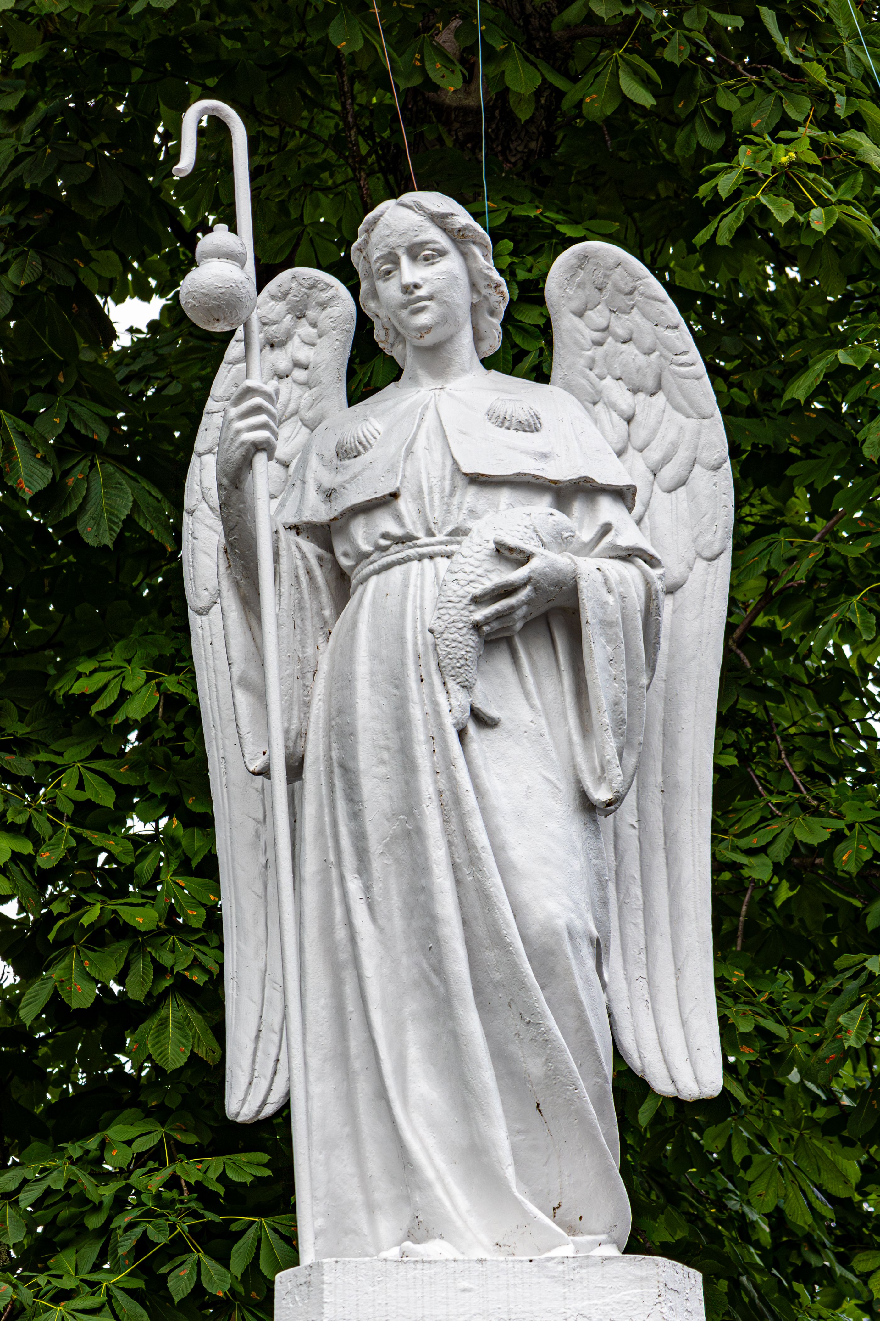 A serene white angel statue stands gracefully against lush green foliage in Lourdes, France.