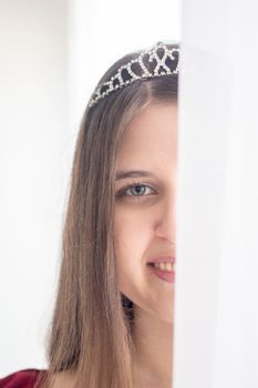 A smiling woman partially hidden by a white curtain, wearing a sparkling tiara indoors.