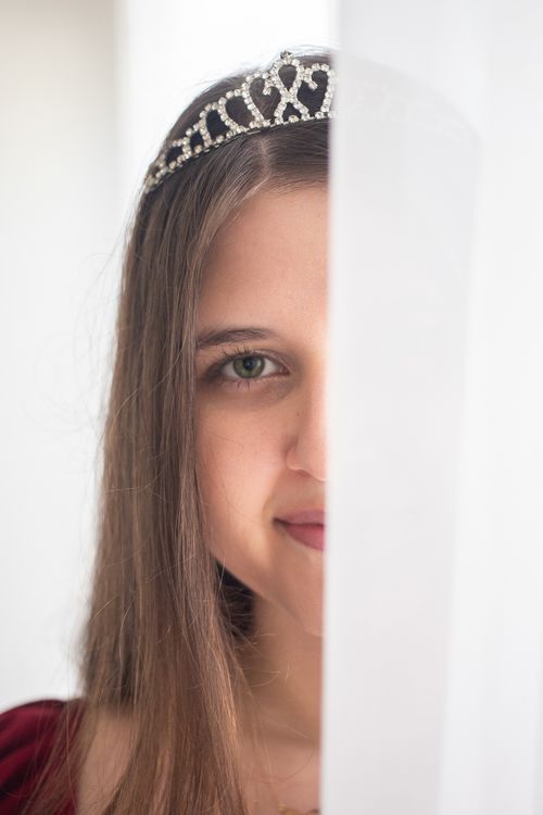 Free A young woman with a tiara peeks behind a curtain, exuding elegance and mystery. Stock Photo