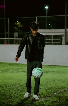 A young man in a leather jacket skillfully playing soccer on an artificial turf field at night.