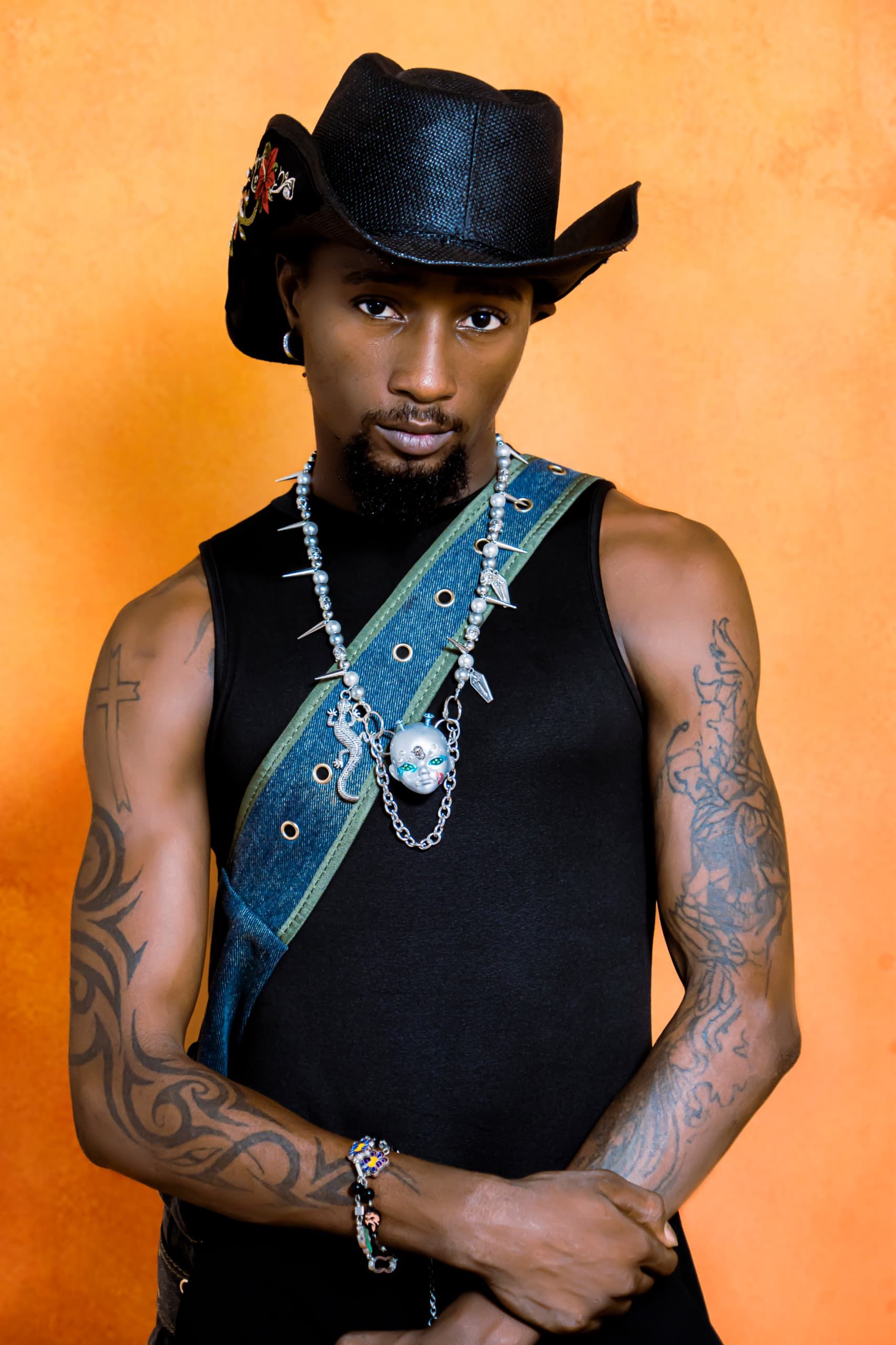 Portrait of a stylish man in a cowboy hat, showcasing tattoos against a vibrant orange background.