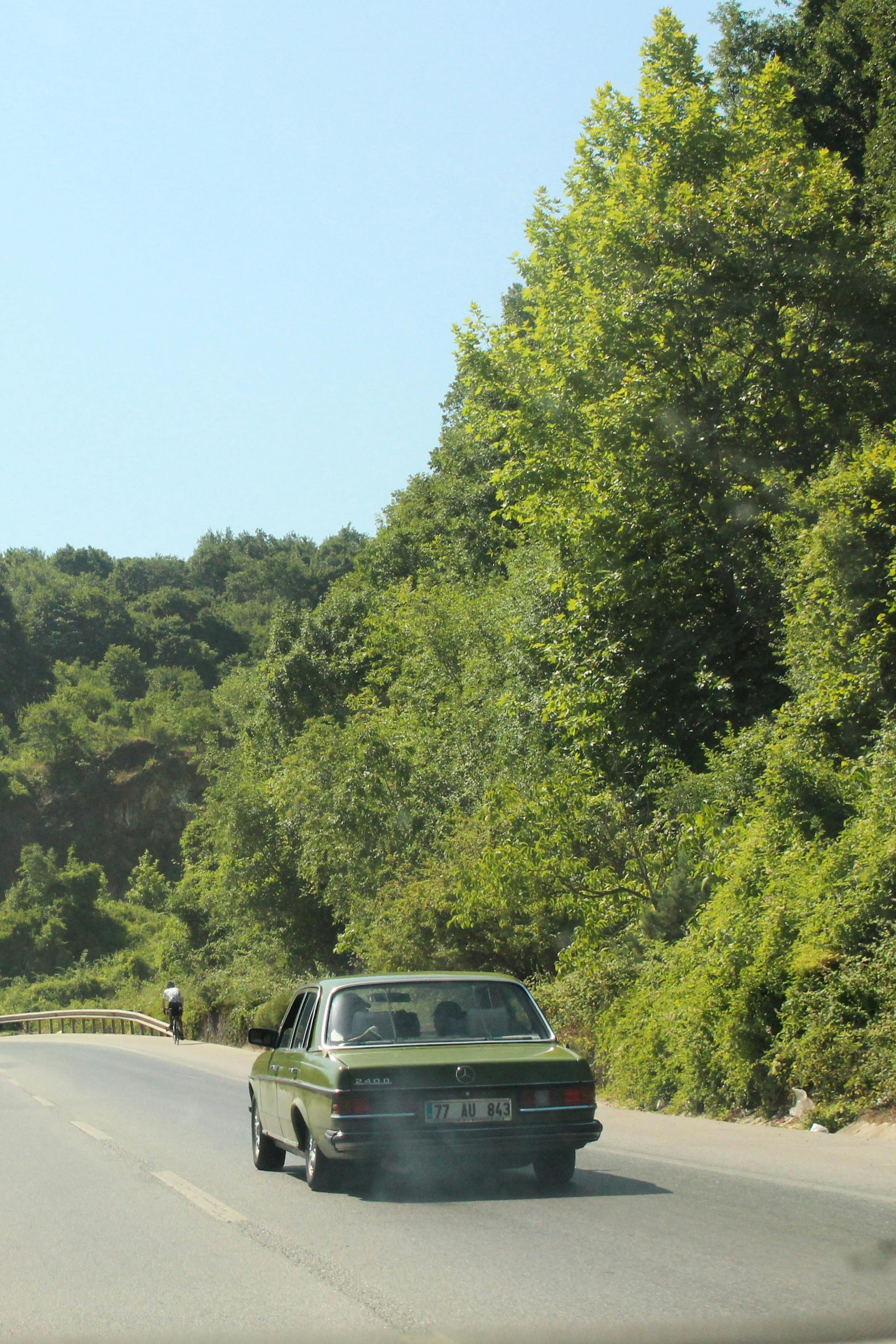 Vintage car driving on a sunny, scenic road surrounded by lush greenery.