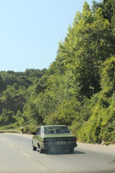 Vintage car driving on a sunny, scenic road surrounded by lush greenery.