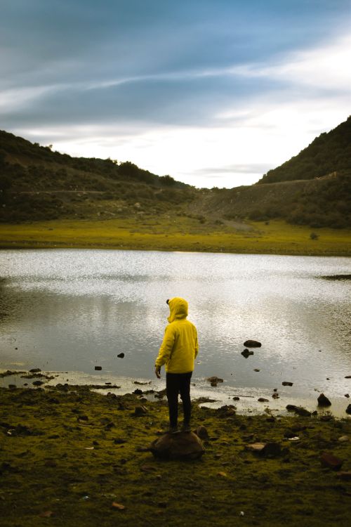 Free A person in a yellow hoodie stands by a tranquil lake surrounded by hills, capturing a peaceful outdoor moment. Stock Photo