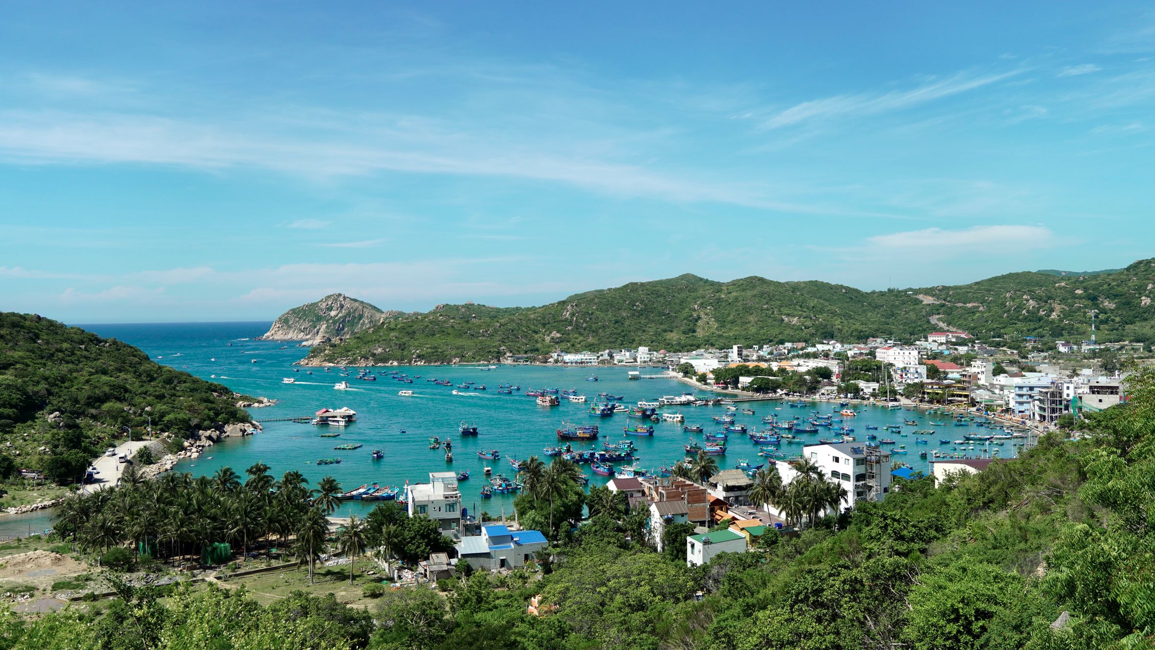 Aerial view of a picturesque fishing village and harbor with boats. Lush greenery and mountains surround the area.