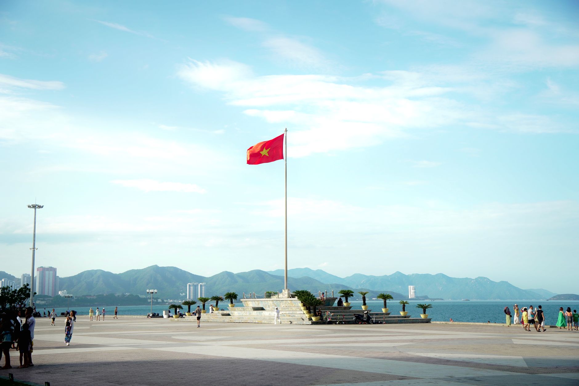 A large Vietnam flag waves along the scenic Nha Trang beachfront with mountains in the background.
