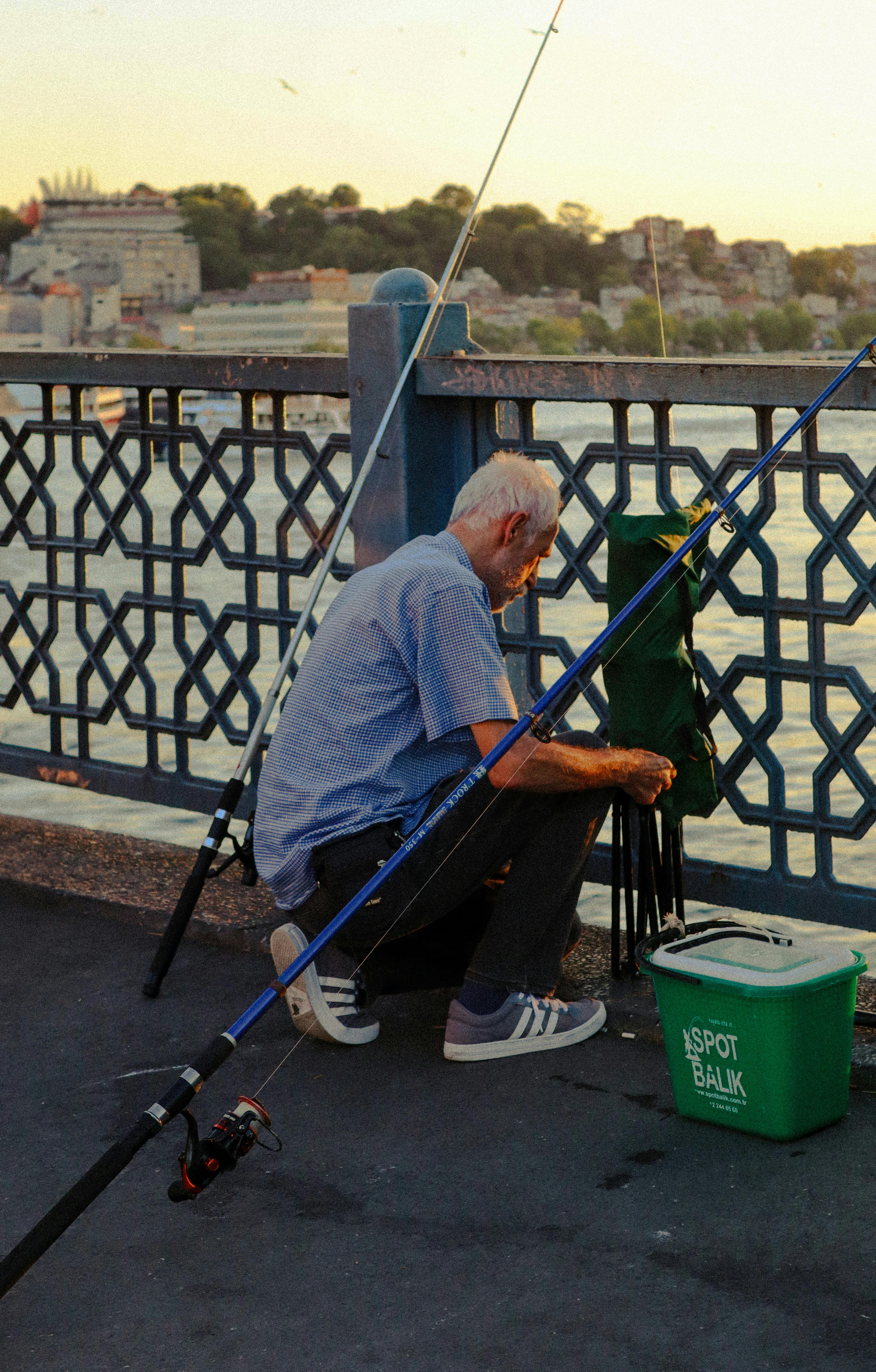 Elderly man fishing on Galata Bridge with sunset view of Istanbul in the background.