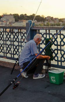 Elderly man fishing on Galata Bridge with sunset view of Istanbul in the background.