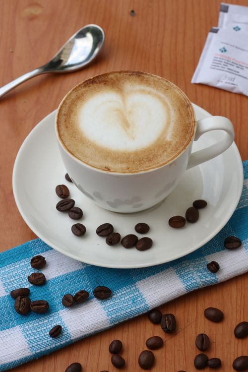 Free A coffee cup with heart-shaped latte art surrounded by coffee beans on a checkered cloth. Stock Photo