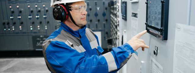 Technician in safety gear working at an industrial control panel indoors.