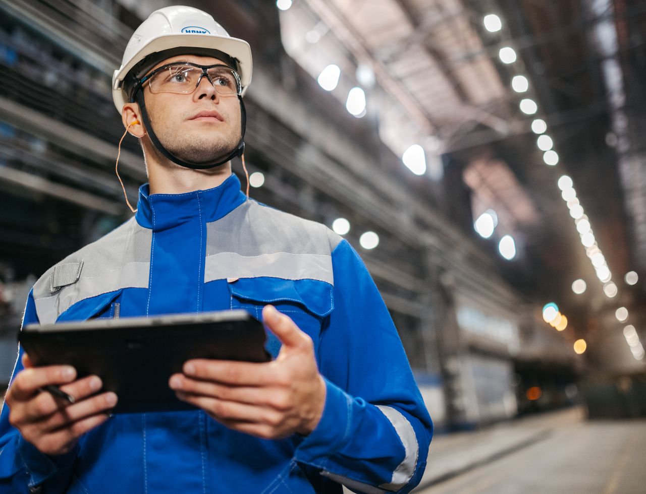 Engineer in a hard hat, safety glasses and work jacket reading a tablet inside a long industrial production hall.