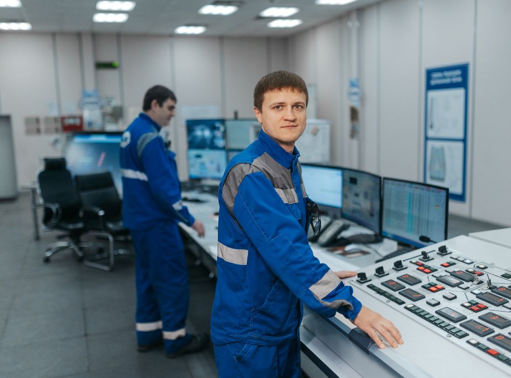 Two technicians in blue coveralls operating a plant control desk with screens and push-button panels.