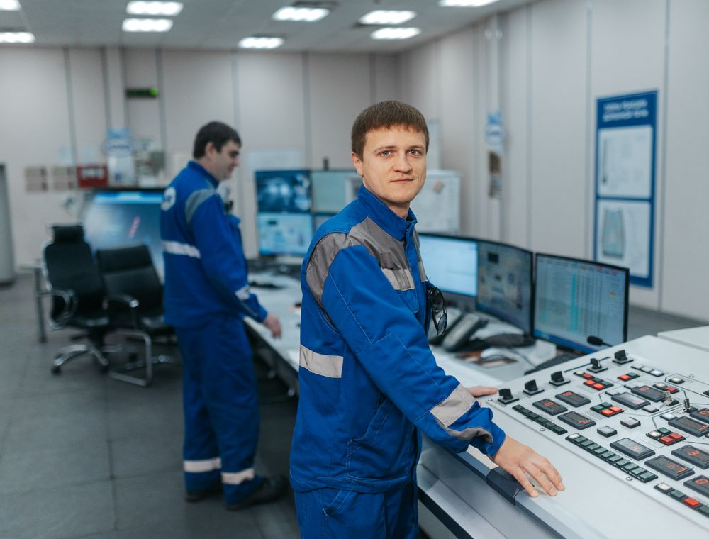 Two technicians in blue coveralls operating a plant control desk with screens and push-button panels.