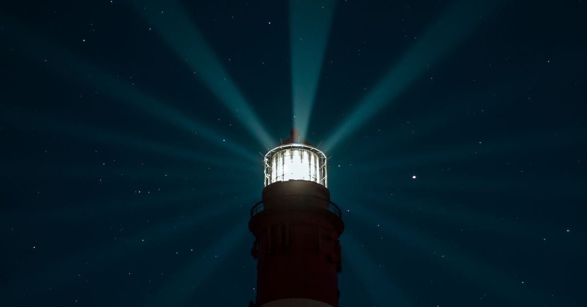 A lighthouse beams light across a starry night sky, creating an ethereal glow over the sea.